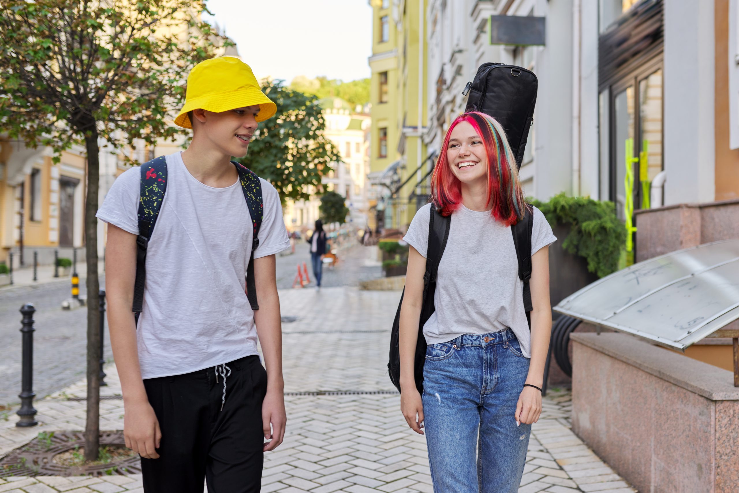 Creative fashionable happy teenagers guy and girl walking and talking along city street with guitar in case. Lifestyle, youth, city life